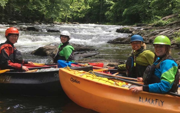 People wearing safety gear sit in canoes and smile at the camera. In the background, there is whitewater and trees lining the shores.